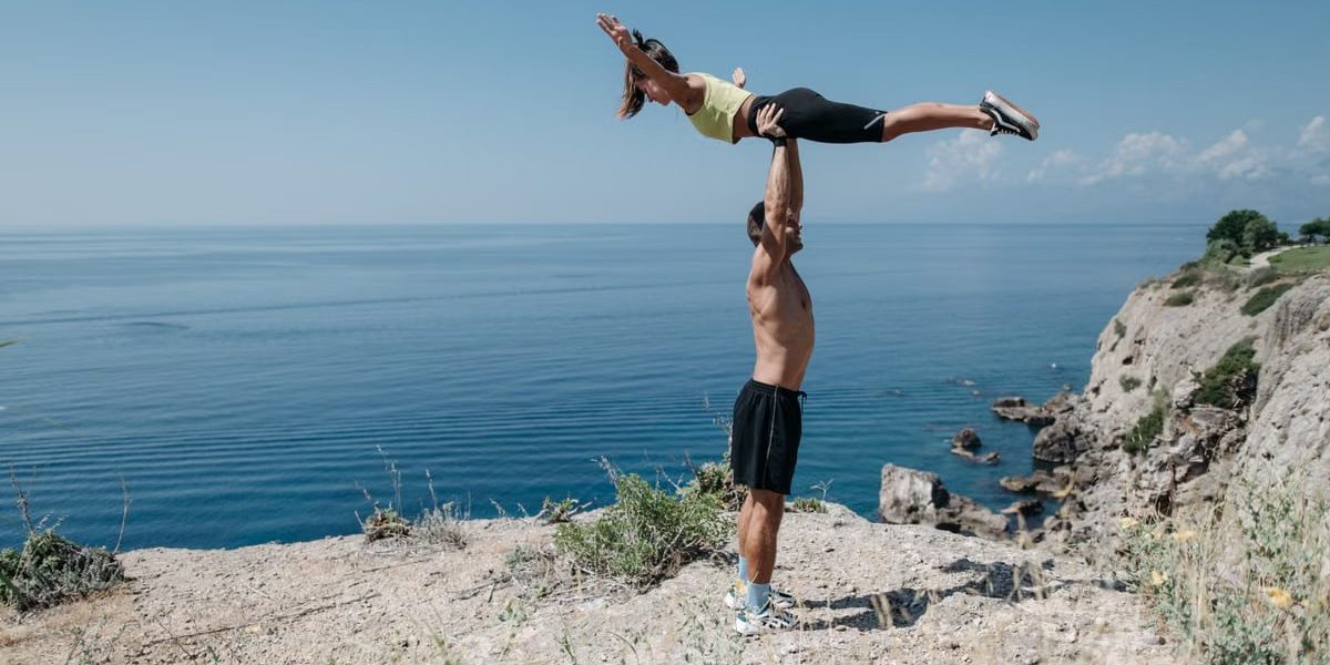 A couple performs acrobatic exercise on a scenic ocean cliff, embodying fitness and adventure.