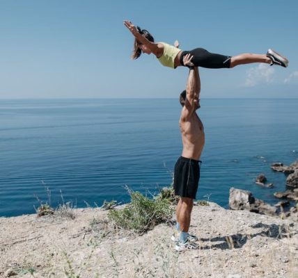 A couple performs acrobatic exercise on a scenic ocean cliff, embodying fitness and adventure.